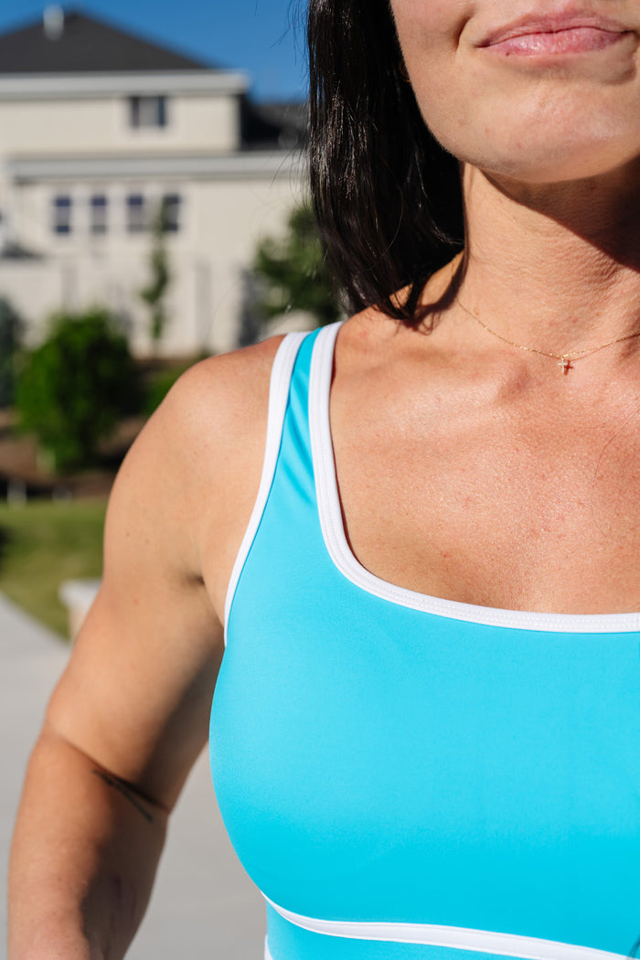 A close-up of a woman wearing a bright turquoise athletic tank top with white trim, photographed outdoors with a blurred building and greenery in the background.
